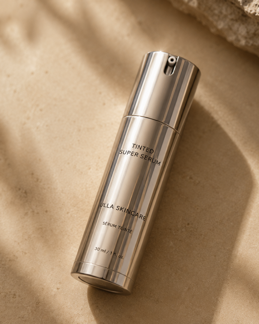 A sleek, metallic silver bottle of Villa Skincare Tinted Super Serum, styled diagonally on a textured beige stone surface with soft leaf shadows cast across it.